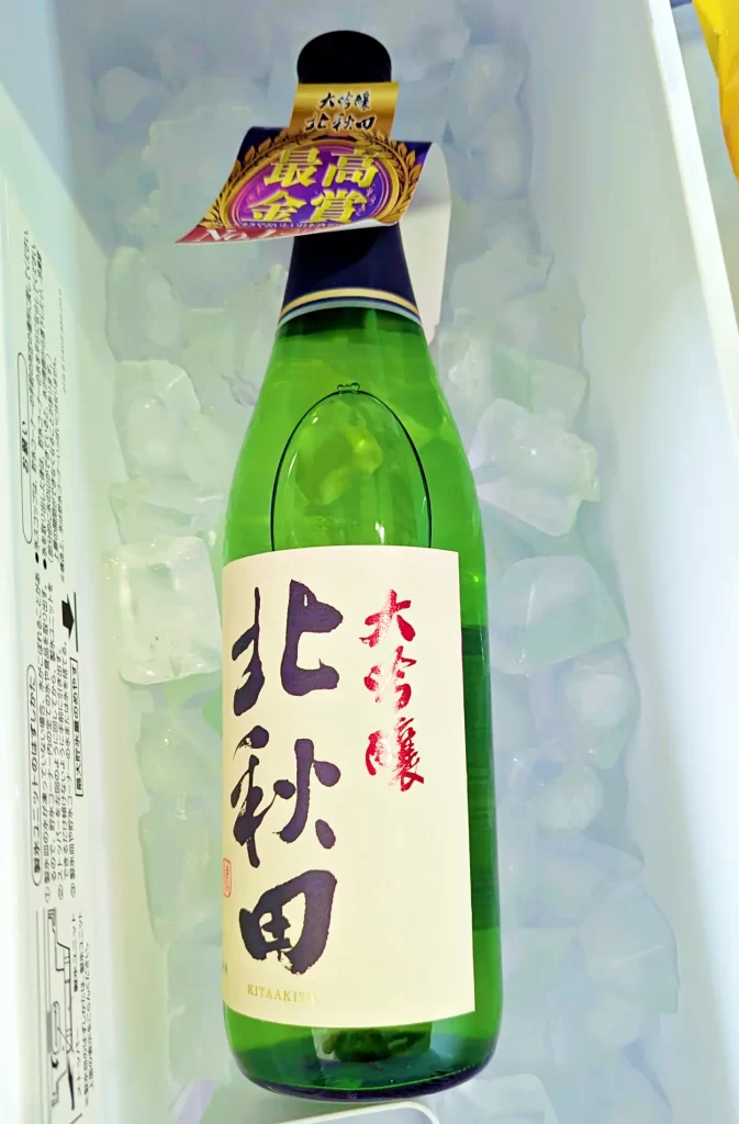 氷で冷やしたクーラーボックスの中に置かれた「大吟醸 北秋田」の四合瓶。緑色のボトルに大きな筆文字のラベルが映え、首元には「最高金賞」の受賞タグが付いている。透明な氷に囲まれた日本酒が、夏の晩酌や冷酒を連想させる爽やかな雰囲気を演出している。