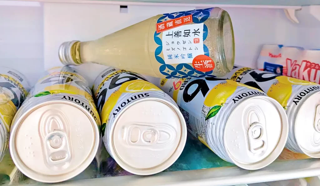 冷蔵庫内で冷やされている「上善如水 純米吟醸 生酒」の300mlボトル。周囲にはレモンサワー缶や乳酸菌飲料が並び、家庭で晩酌用に保存されている様子がうかがえる。瓶には冷蔵推奨の表示があり、表面には結露した水滴が付いている。